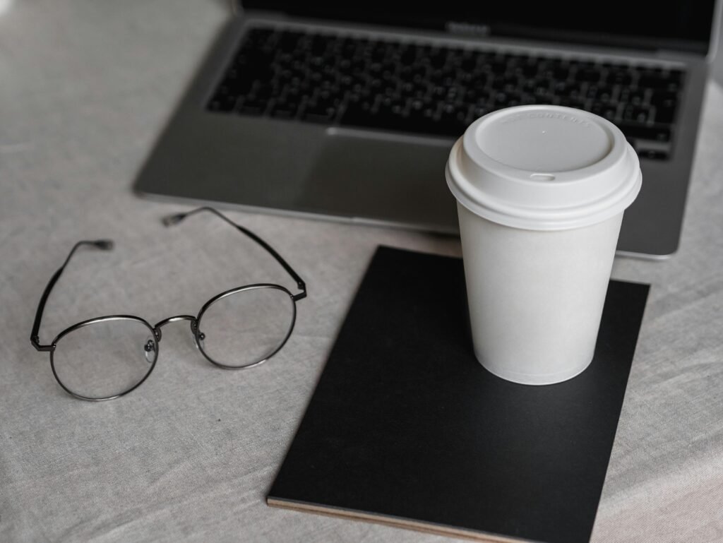 Disposable Cup on Black Notebook Beside a Laptop and Eyeglasses